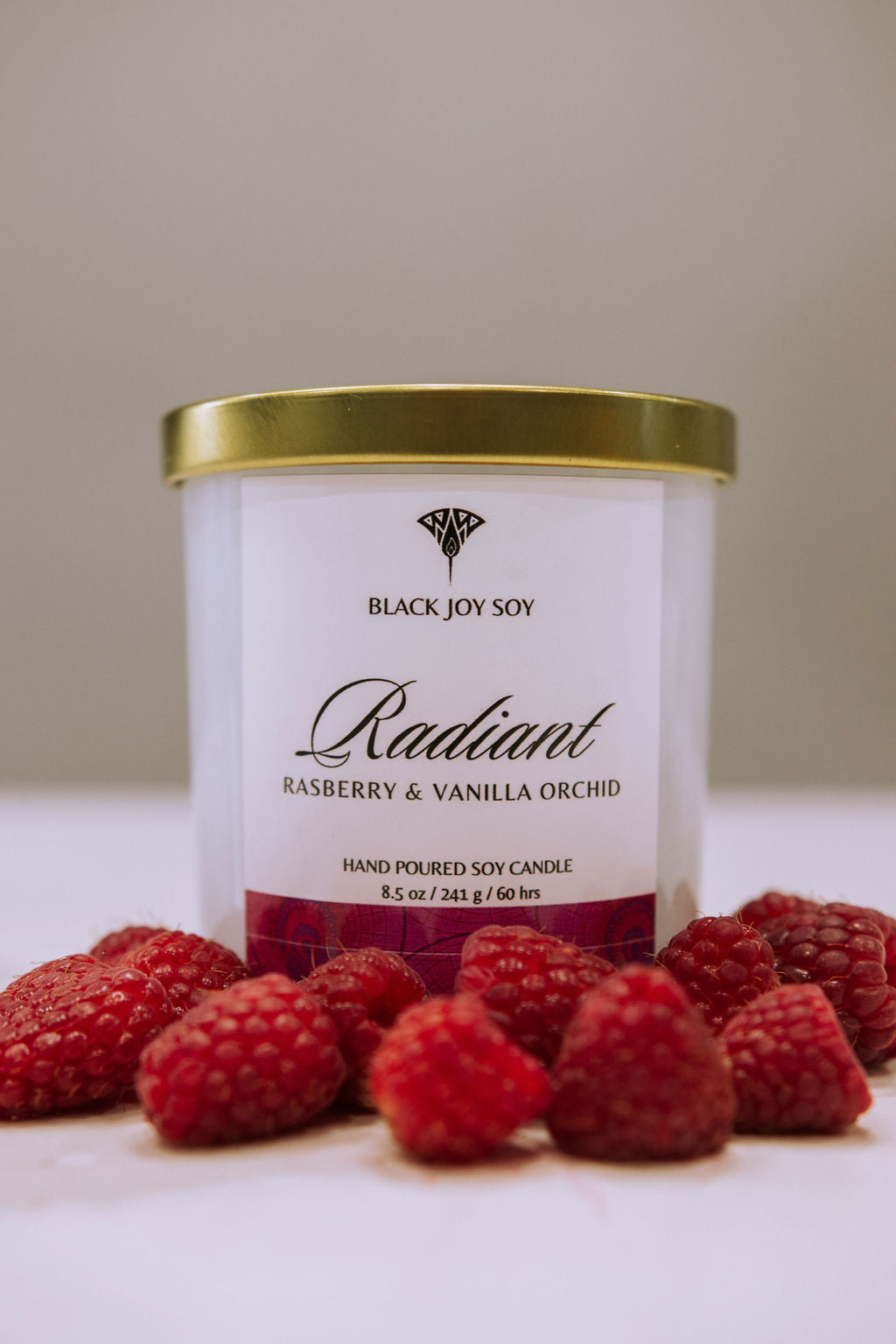 This is a 8.5 ounce Raspberry, Vanilla & Orchid scented soy candle named "Radiant" in a white jar with a gold lid, and a bold pink label. There are fresh raspberries surrounding the candle jar.