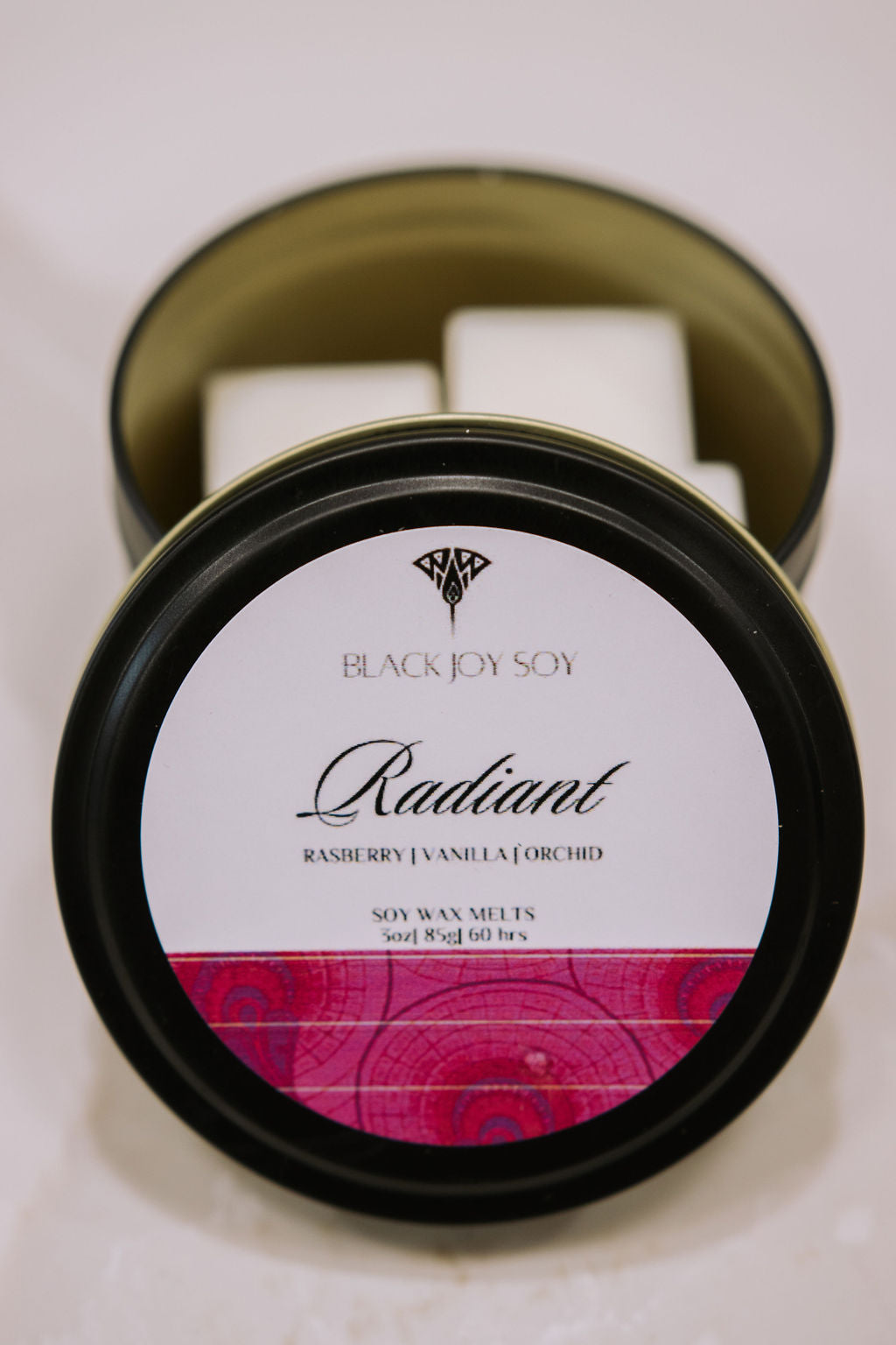 black metal tin with 16 Raspberry, Vanilla & orchid scented soy wax melts inside. the tin lid has a hot pink label on it with the word "Radiant" on it.