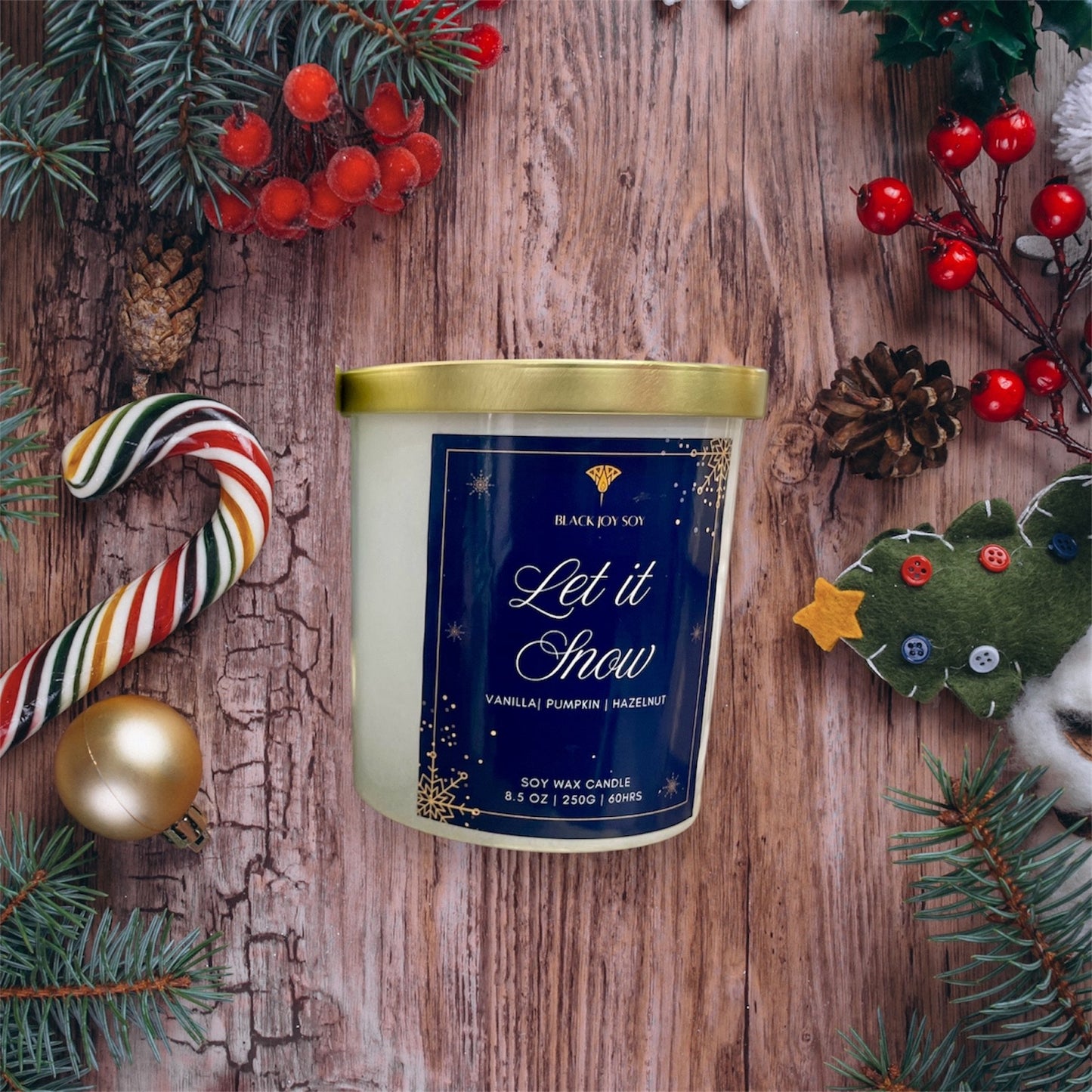 Candle labeled 'Let it Snow' surrounded by Christmas decorations on a wooden surface. vanilla, pumpkin and hazelnut scented soy candle