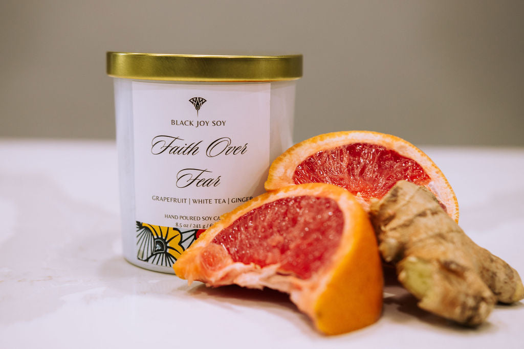 This is a 8.5 ounce Grapefruit, white tea & ginger soy scented candle named "Faith Over" in a white jar with a gold lid, and a red, yellow and white label. There is fresh grapefruit and ginger depicted next to the candle.