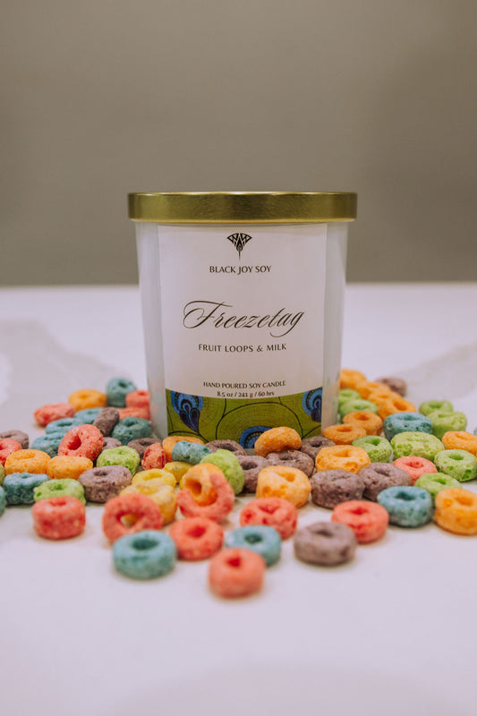 This is an 8.5 ounce fruit loops soy scented candle named "Freezetag" in a white jar with a gold lid, and bright green label with blue accents. There are fruit loops surrounding the candle jar.
