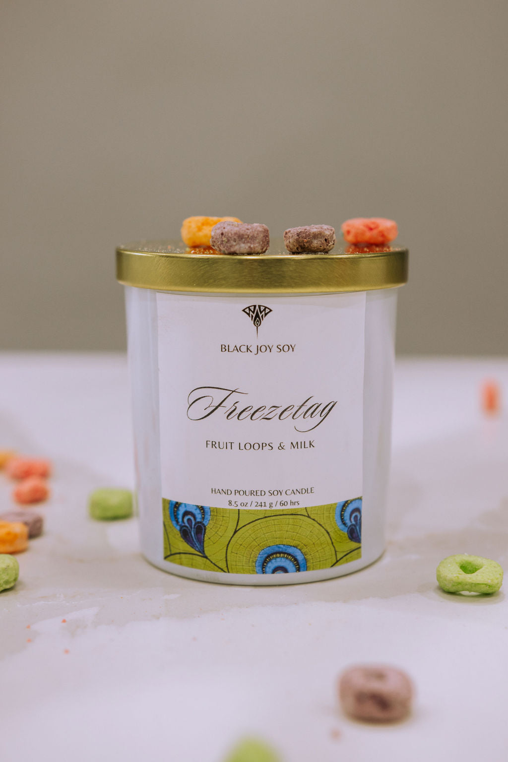 This is an 8.5 ounce fruit loops scented soy candle named "Freezetag" in a white jar with a gold lid, and bright green label with blue accents. There are fruit loops surrounding the candle jar.