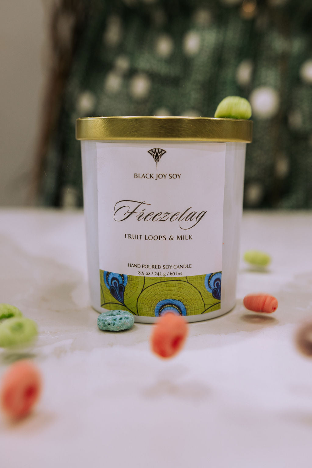 This is an 8.5 ounce fruit loops scented soy candle named "Freezetag" in a white jar with a gold lid, and bright green label with blue accents. There are fruit loops surrounding the candle jar.