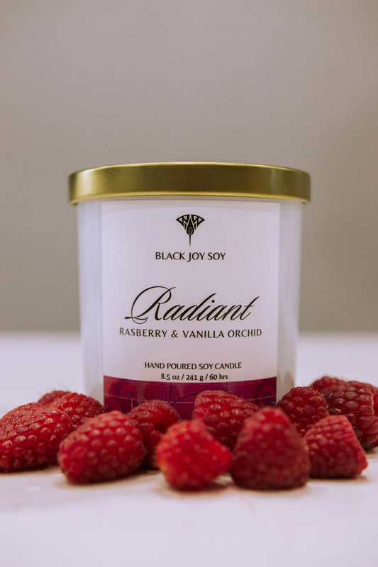 This is a 8.5 ounce Raspberry, Vanilla & Orchid scented soy candle named "Radiant" in a white jar with a gold lid, and a bold pink label.  There are fresh raspberries surrounding the candle jar.