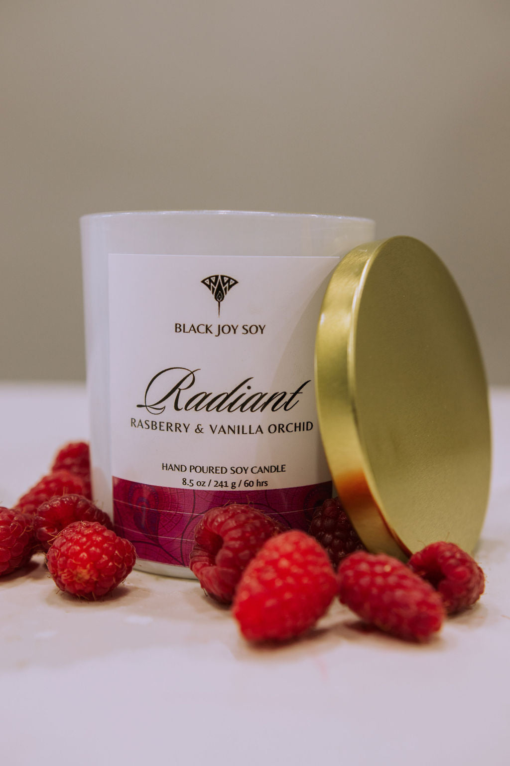 This is a 8.5 ounce Raspberry, Vanilla & Orchid scented soy candle named "Radiant" in a white jar with a gold lid leaned against the jar and a bold pink label.   There are fresh raspberries surrounding the candle jar.