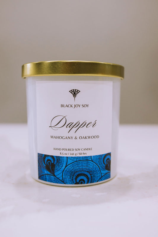 This is a 8.5 ounce Mahogany & Teakwood scented candle named "Dapper" in a white jar with a gold lid, and a bold royal blue label. 