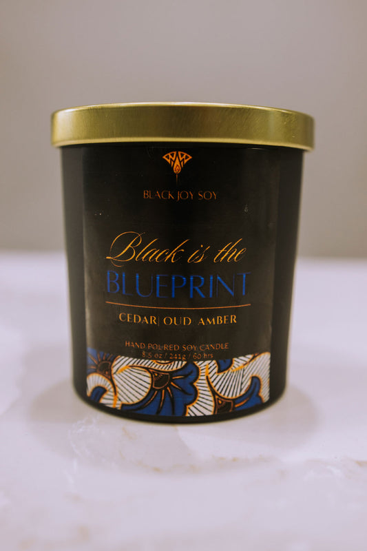 This is an 8.5 ounce amber, red cedar and oud scented candle named "Black is the Blueprint" in a black jar, with a gold lid, and a black, royal blue and gold label.