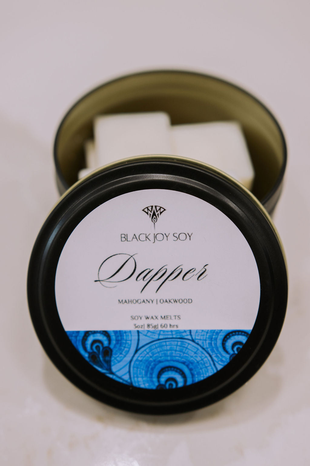 black metal tin with 16 Mahogany & Teakwood scented soy wax melts inside. the tin lid has a blue label on it with the word "Dapper" on it.
