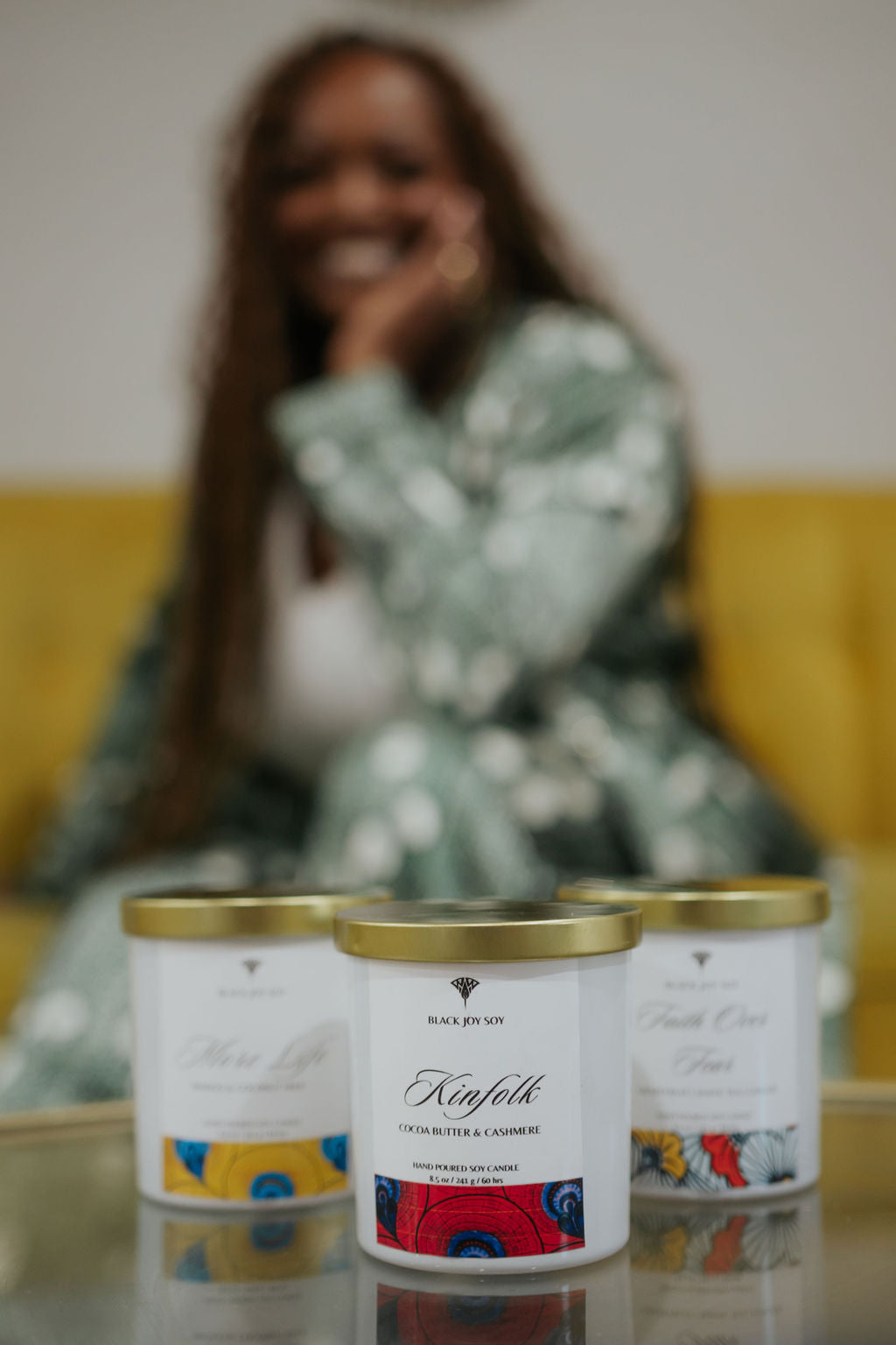 Cocoa butter cashmere soy candle depicted in foreground. The candle is in a white jar with a bold red label that has dark blue accents. A blurred image of a smiling woman  (the company founder) is in the background. 