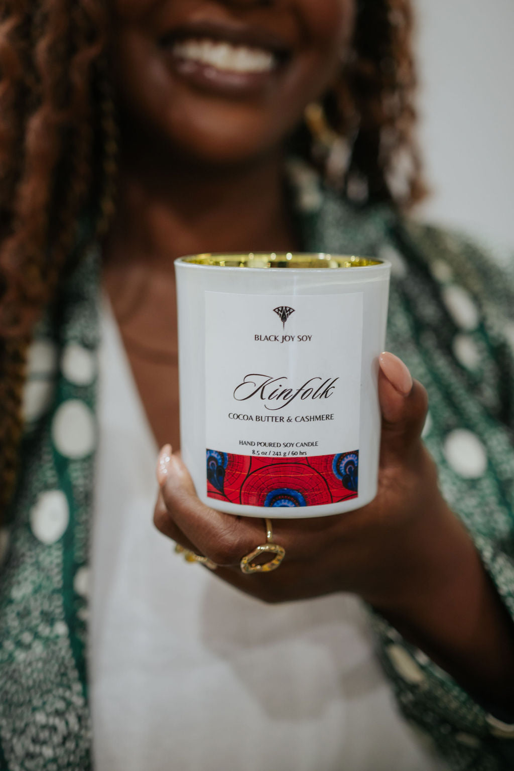 Cocoa butter cashmere soy candle depicted in foreground. Candle is being held by a smiling woman who is in the background. The candle is in a white jar with a bold red label that has dark blue accents.