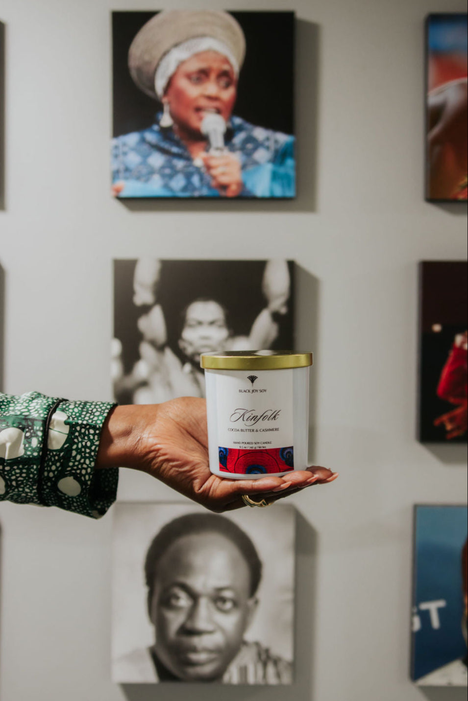 Cocoa butter cashmere soy candle depicted in foreground. Candle is being held in front  of pictures of various Black historic figures. The candle is in a white jar with a gold lid and a bold red label that has dark blue accents.