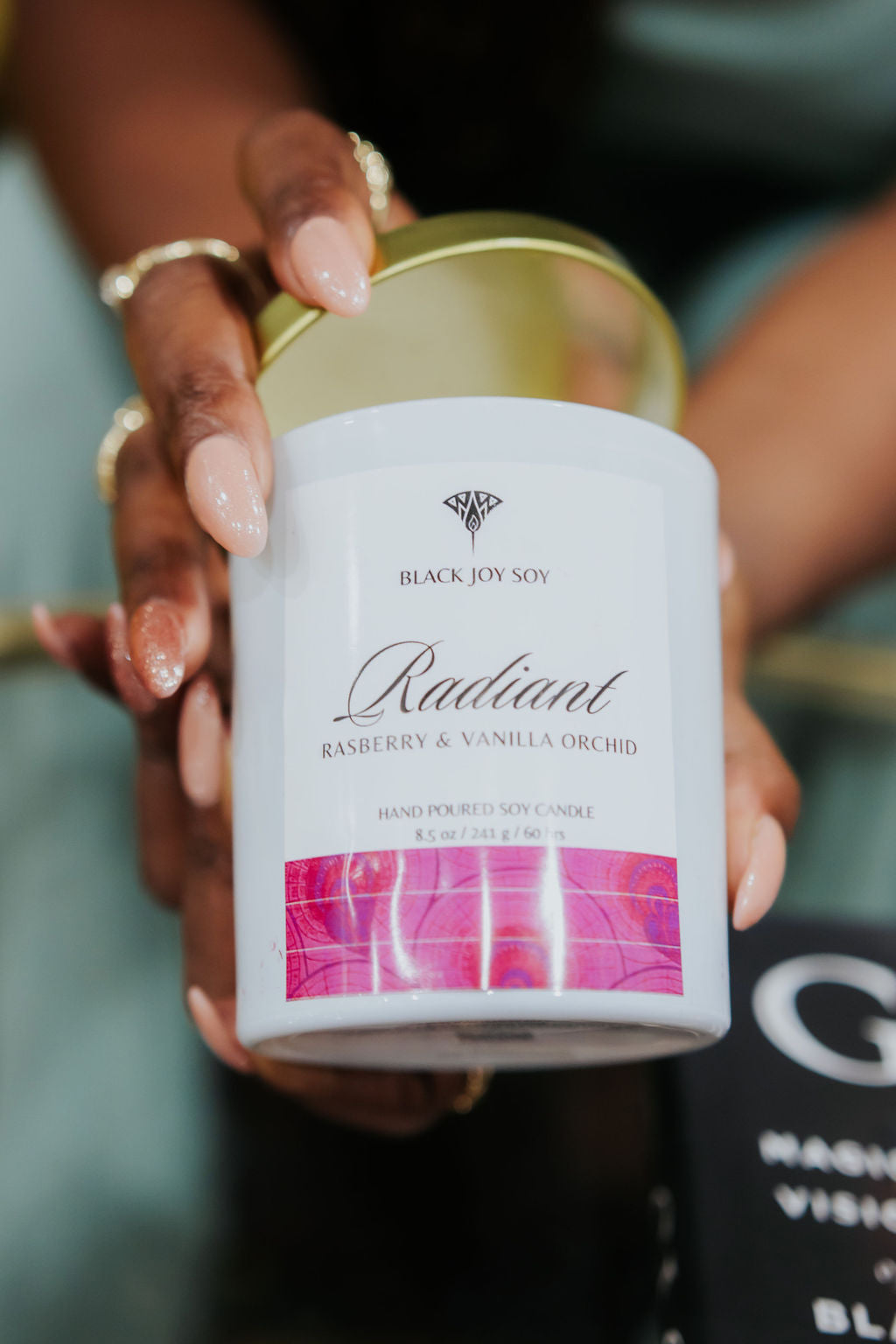 The hands of a woman are holding a raspberry vanilla soy candle in a white jar with a gold lid and a pink and white label that says "Radiant".