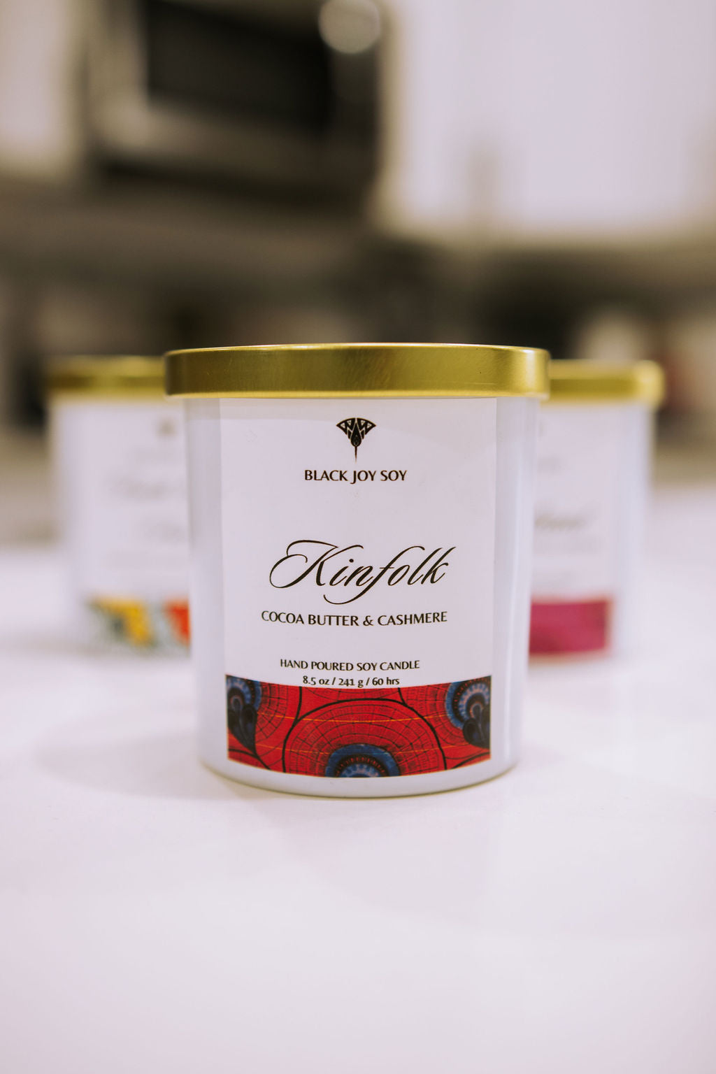 This is a 8.5 ounce Cocoa Butter & Cashmere scented soy candle named "Kinfolk" in a white jar with a gold lid, and a bold red label with royal blue accents. 