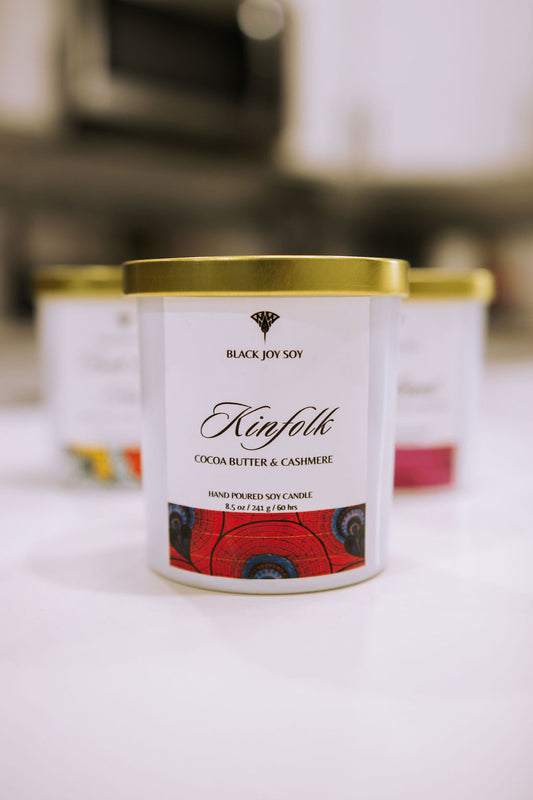 This is a 8.5 ounce Cocoa Butter & Cashmere scented soy candle named "Kinfolk" in a white jar with a gold lid, and a bold red label with royal blue accents. 