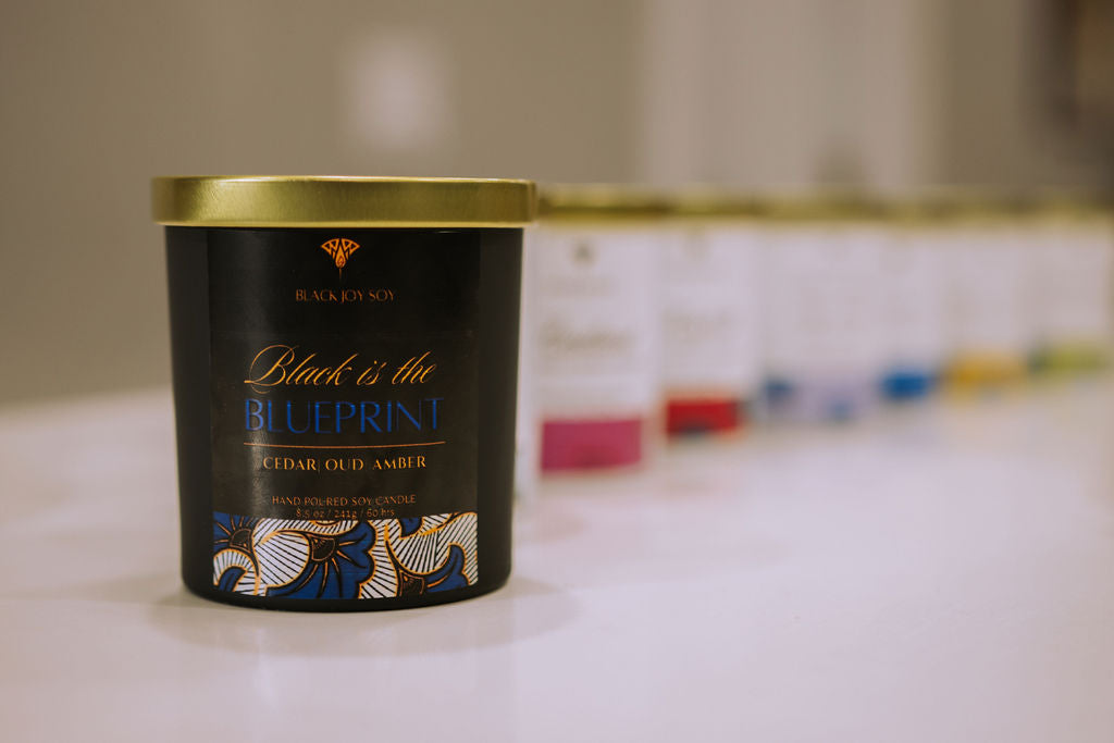 This is an 8.5 ounce amber, red cedar and oud scented candle named "Black is the Blueprint" in a black jar, with a gold lid, and a black, royal blue and gold label.
