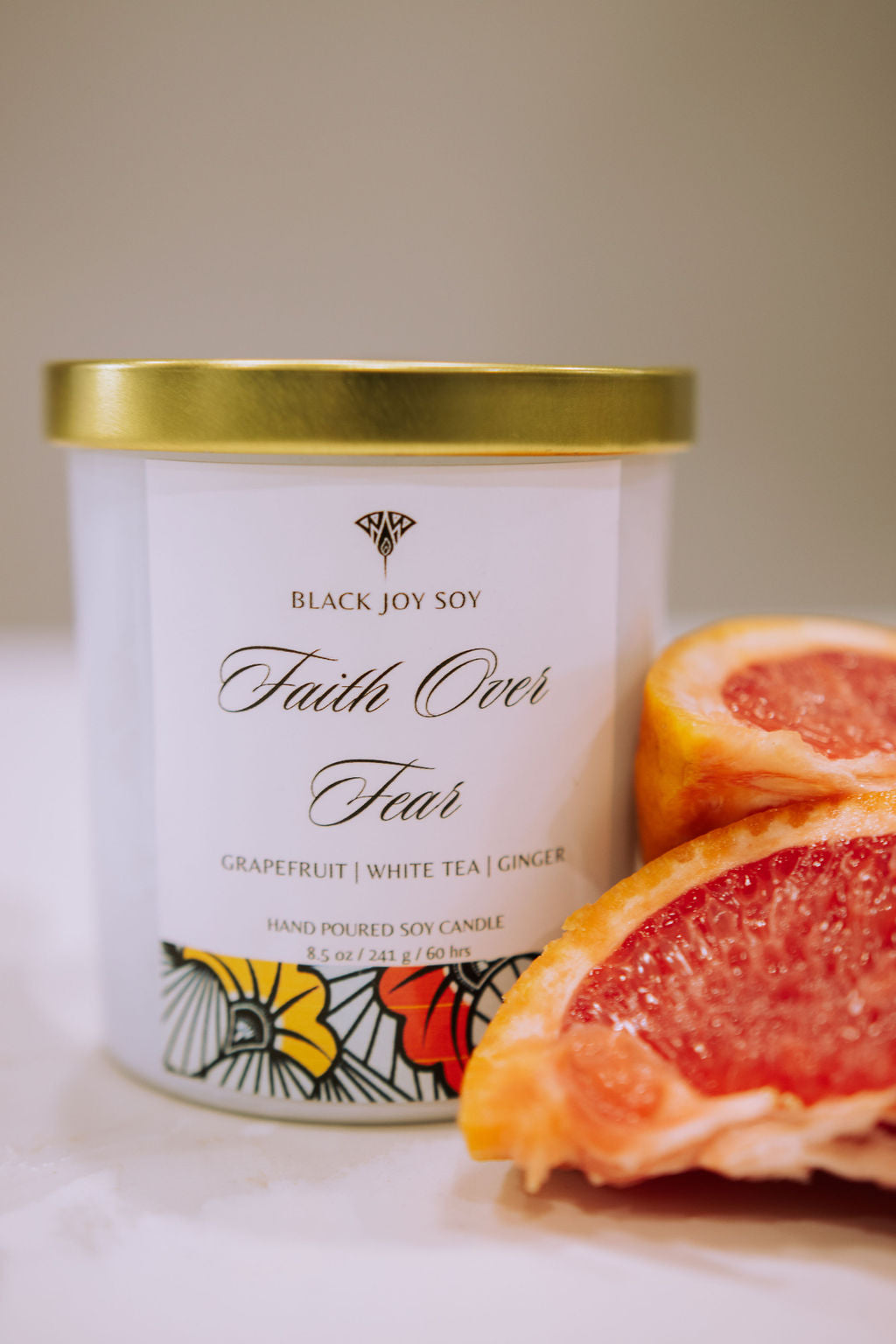 This is a 8.5 ounce Grapefruit, white tea & ginger scented soy candle named "Faith Over" in a white jar with a gold lid, and a red, yellow and white label. There is fresh grapefruit depicted next to the candle.
